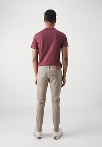 Camiseta de algodón color granate combinada con vaqueros ajustados beige claro. Costuras notables en el bolsillo trasero y parche de cuero con la marca. Zapatillas blancas completan el conjunto.