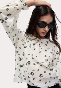 Camicia bianca a maniche lunghe con motivo floreale nero, vestibilità ampia e tessuto testurizzato. La modella indossa occhiali da sole rettangolari neri.