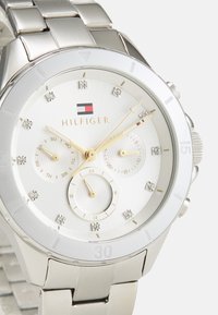 Montre en acier inoxydable argenté avec un cadran blanc, des aiguilles dorées et des accents en cristal. Dispose de trois sous-cadrans et d'une lunette polie.