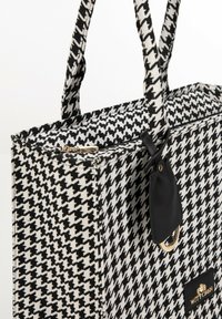 Sac fourre-tout à motif pied de poule noir et blanc fabriqué en tissu, doté de deux poignées tressées, d'une fermeture éclair et d'un accent détachable noir.