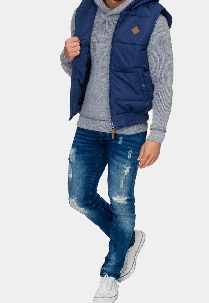 Homme portant une veste matelassée bleue par-dessus un pull en maille gris, un jean bleu déchiré et des baskets blanches, debout avec une jambe croisée.