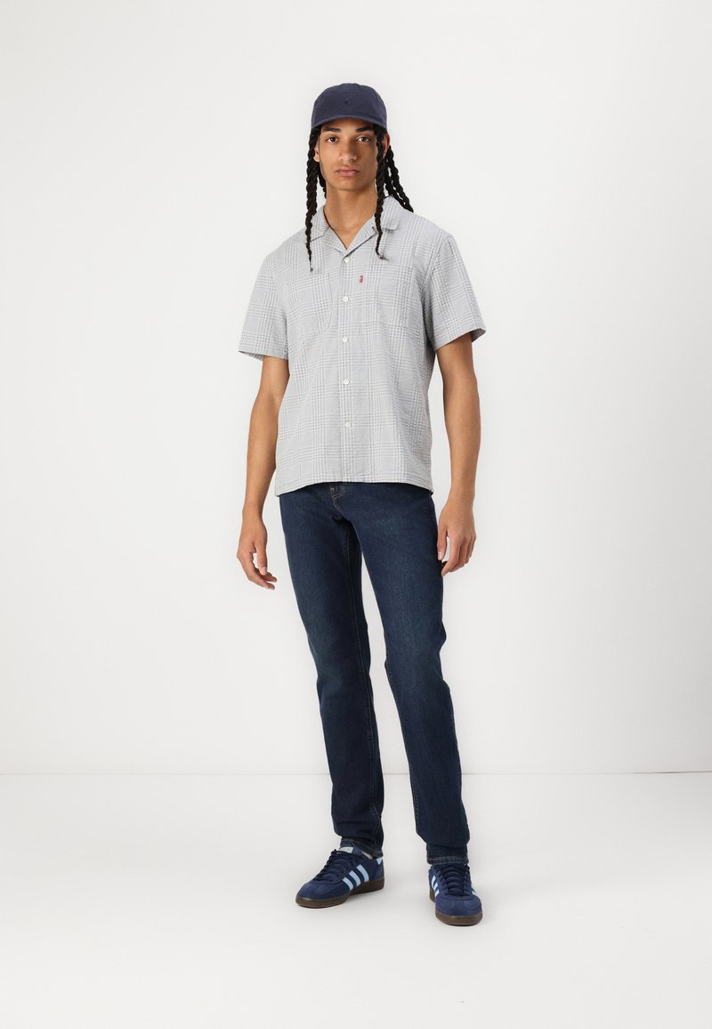 Chemise à manches courtes à carreaux gris clair ; jean bleu foncé ; baskets bleues avec des rayures bleu clair ; casquette marine.