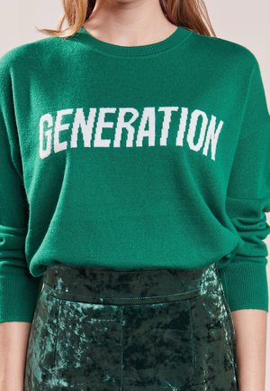 Osoba nosząca zielony sweter z napisem "GENERATION" białymi literami oraz zieloną aksamitną spódnicę.