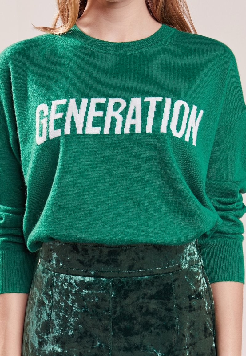 Personne portant un pull vert avec « GENERATION » en lettres blanches et une jupe verte en velours.