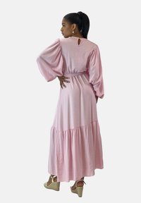 Vestido largo rosa con mangas abullonadas, detalle de lazo en la parte trasera y falda de capas, hecho de tela ligera. Se lleva con sandalias de cuña.