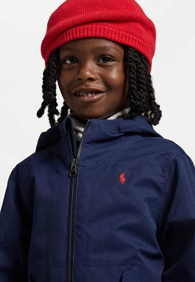 Temno modra jakna z zadrgo spredaj, ki ima majhen rdeč logotip na prsih; v kompletu s svetlo rdečim pletenim kapucarjem.