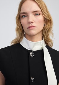 Czarna sukienka bez rękawów z białym jedwabnym kołnierzykiem i krawatem, zdobiona srebrnymi broszkami oraz fakturowymi akcentami wokół dekoltu.