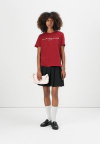 Raudona medvilninė marškinė su balta "TOMMY HILFIGER" logotipu, derinama su juoda klostuota sijonu, baltais kelio ilgio kojiniais ir šviesiai rožine rankine.