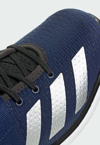 Modré sportovní boty s texturovaným síťovaným svrškem, černými tkaničkami a třemi výraznými bílými pruhy po straně. Viditelná gumová podrážka.