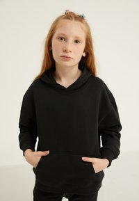 Melnā kapuce ar priekšējo kabatu, brīvas formas un ar kapuci. Izgatavota no mīksta auduma ar ribotām aprocēm. Modelis ar garām sarkanām matiem un pieres plankumiem.