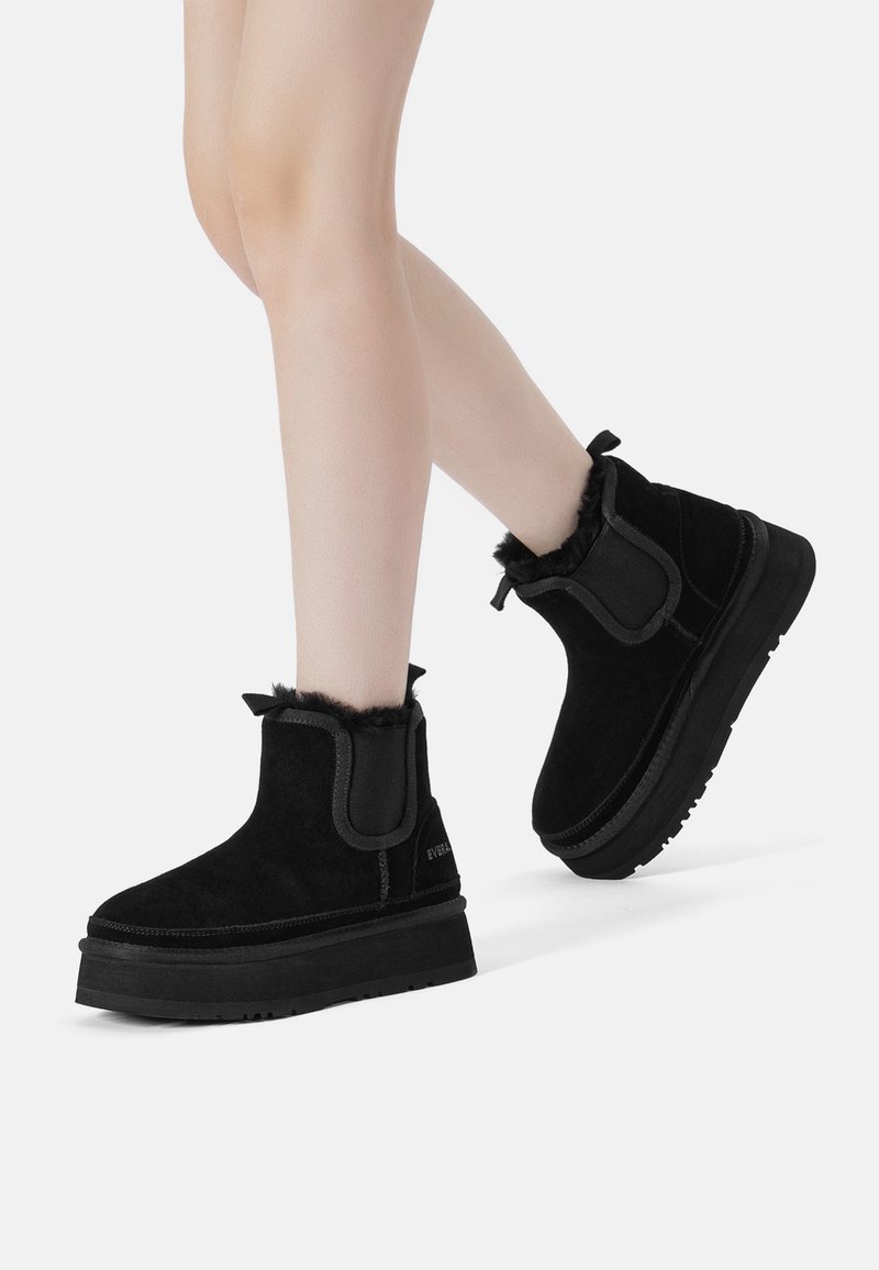 Black ankle boots made of suede, featuring a faux fur trim, elastic side panels, and a thick rubber sole with textured tread.