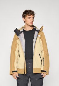 Bež jaqueta ja rainproof, mis on varustatud kapuutsiga, lukuga taskutega, reguleeritavate mansettidega ja võrkkattega voodriga. Kantud musta topi peal.