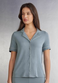 Krajkové pyžamové tričko s krátkým rukávem, vyrobené z jemné modré látky, s špičatým límečkem a bílým lemováním po okrajích. Hladká textura.