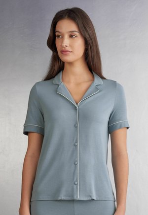 Kortaf, knoopsluiting pyjamatok in een zachte blauwe stof, met een puntcollar en witte piping langs de randen. Soepele textuur.