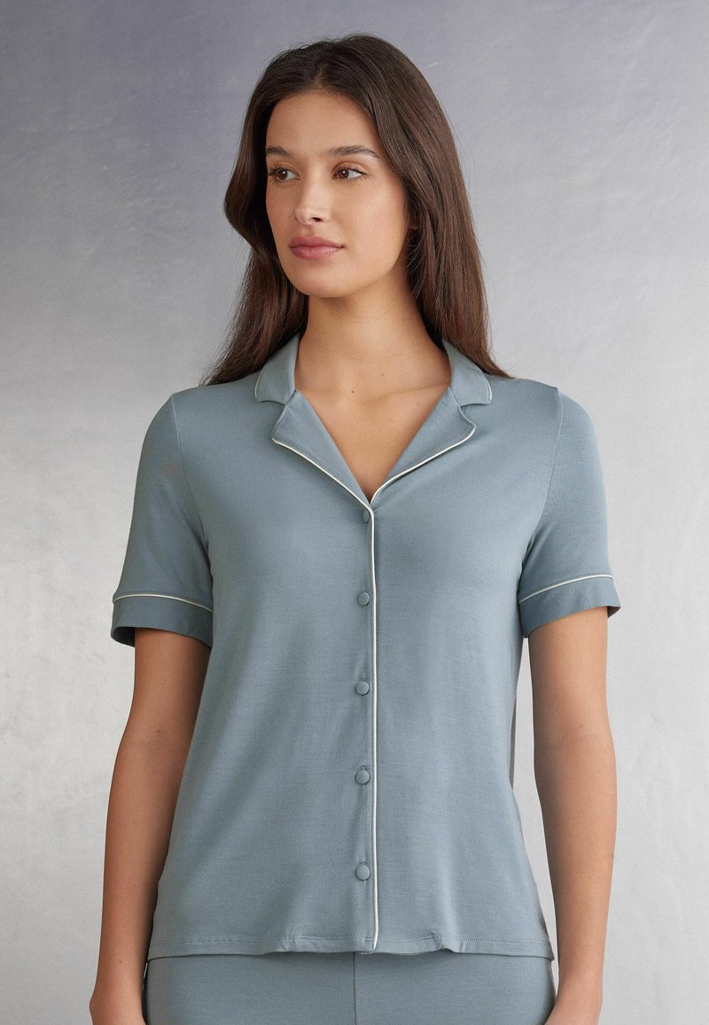 Krajkové pyžamové tričko s krátkým rukávem, vyrobené z jemné modré látky, s špičatým límečkem a bílým lemováním po okrajích. Hladká textura.