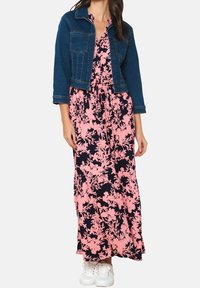 Jeansjacka över en blommig maxiklänning med rosa blommor på marinblå bakgrund, med insvängd midja och vita sneakers.