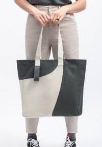 Canvas totebag in beige en donker groen, met een geometrisch ontwerp, twee stevige handvatten en een label met een grafisch logo.