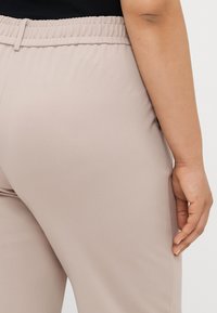 Lys beige bukser med elastisk talje. Glat, strækbart stof, tætsiddende design, viser en del af en hånd hvilende på hoften.