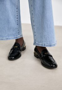 Mocasines de charol negro con un acento dorado, que presentan un logo de GUESS en la correa. Combinados con unos vaqueros de denim azul claro de pierna ancha.