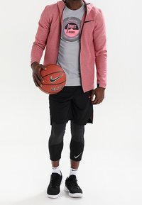 Roter sportlicher Hoodie, graues T-Shirt mit Grafik, schwarze Shorts, schwarze Leggings und schwarze Basketballschuhe, hält einen Basketball in der Hand.
