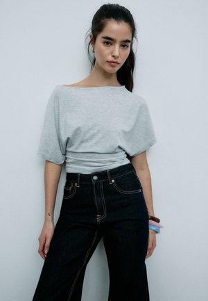 Ung kvinde med mørkt hår sat op i en hestehale iført en løs lysegrå top og højtaljede mørke jeans, stående op ad en ensfarvet væg.