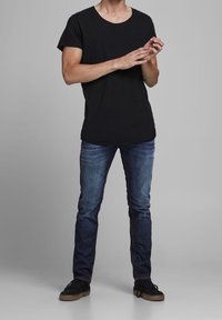 Camiseta negra de manga corta con escote redondo, combinada con jeans de mezclilla azules. Zapatillas negras completan el atuendo, con un fondo gris.