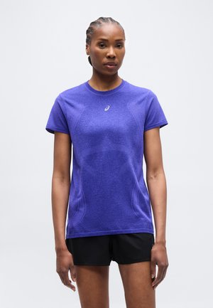 Kvinne med flettehår iført lilla kortermet treningsskjorte og svarte shorts, stående foran en ensfarget lys bakgrunn.