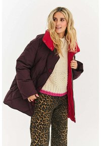 Pikowana kurtka puchowa w ciemnym bordowym kolorze z jasnoczerwonym kołnierzem, wyposażona w duże guziki i oversize'owy krój. Noszona na swetrze dzianinowym i spodniach w panterkę.