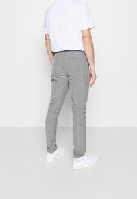 Muž v šedých kostkovaných kalhotách, bílých teniskách a bílém tričku, stojící zády k fotoaparátu na jednobarevném pozadí.