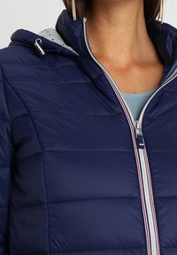 Chaqueta acolchada azul marino con cremallera frontal, con líneas acolchadas horizontales y forro de capucha gris. Incluye una cremallera plateada con detalles en rojo.