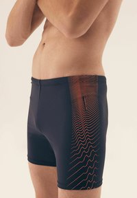 Granatowe kąpielówki z teksturowanym pomarańczowym falistym wzorem po boku, wykonane z elastycznej tkaniny zapewniającej komfort i swobodę ruchów.