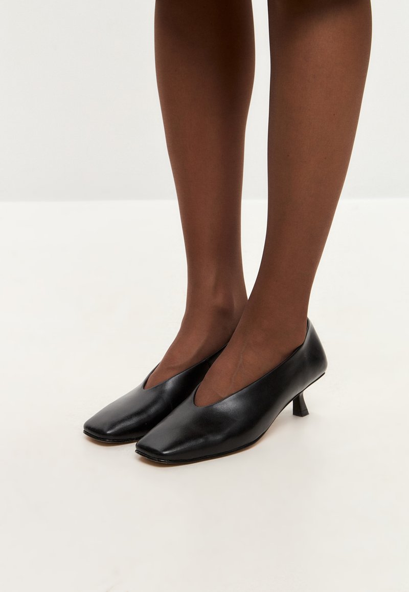 Scarpe con tacco alto in pelle nera, con punta quadrata e design a V, appoggiate su uno sfondo chiaro, indossate con calze trasparenti marroni.