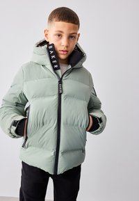 Jonge jongen draagt een lichtgroene winterjas met capuchon, handen in de zakken, kijkt recht in de camera tegen een effen achtergrond.
