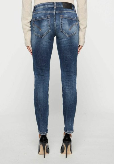 Des jeans en denim bleu slim présentent une taille mi-haute, des poches arrière avec des détails de couture et un léger délavage. Portés avec des chaussures à talons hauts noires.