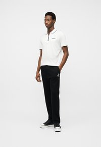 Jeune homme debout portant un polo blanc à manches courtes avec fermeture éclair et un pantalon de jogging noir avec un graphisme blanc sur la poche, baskets noires.
