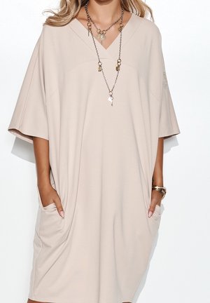 Robe beige oversize avec col en V, manches courtes, poches latérales et texture lisse. Accentuée par un collier à plusieurs rangs avec des breloques.