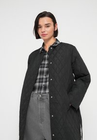 Μαύρο καπιτονέ παλτό με κουμπώματα, με σχέδιο ρόμβου, συνδυασμένο με μαύρο και άσπρο πουκάμισο καρό και γκρι ψηλόμεσα τζιν.