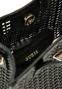 Sac à main tissé noir avec un intérieur lisse, doté d'une poche zippée et de ferrures dorées. Logo "GUESS" embossé à l'intérieur.