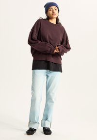 Felpa oversize color marsala indossata sopra una maglietta nera, abbinata a jeans azzurri e scarpe nere con dettagli bianchi. Il berretto è blu navy.