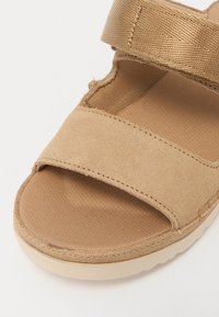 Beige semsket sandal med to brede, myke stropper. Teksturert såle og lys gummisåle. Synlige sømmer langs kantene.