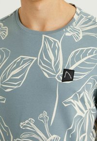 Vaaleansininen puuvillapaita, jossa on suuri valkoinen kasvituloste, pyöreä kaula-aukko ja pieni musta neliölabeli logolla rintataskussa.