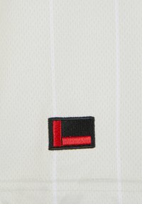 Un tissu blanc avec un tissage texturé présente un logo rectangulaire brodé noir et rouge dans la partie inférieure. Des rayures verticales blanches sont visibles.