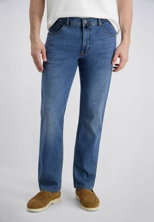 Man draagt mediumblauwe rechte jeans, bruine suède instapschoenen en een wit overhemd, staand tegen een effen achtergrond.