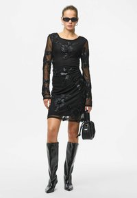 Kobieta ubrana w czarną, przezroczystą sukienkę z długim rękawem, zdobioną kwiatowym haftem, czarne błyszczące kozaki do kolan, czarną torebkę oraz ciemne okulary przeciwsłoneczne.