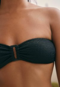 Svart bandeau-bikinitopp med texturerad tyg, volangkanter och en mittknäppning. Slät, sömlös design utan axelremmar.