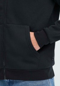 Svart zip-up hettegenser med frontlommer, ribbestrikkede mansjetter og nederkant. Myk, teksturert stoff synlig, med en minimal sømdetalj på siden.