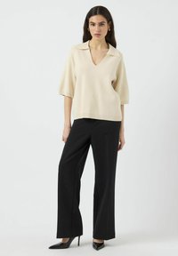 Beige neulotut pusero, jossa on V-mallinen kaula-aukko ja leveät lyhyet hihat, yhdistettynä mustiin leveisiin housuihin ja mustiin kärkitossuihin.