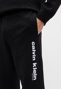 Juodi treningai, pagaminti iš medvilnės mišinio, su šonine kišenė ir baltais užrašais "calvin klein" ir "New York City" ant kojos.