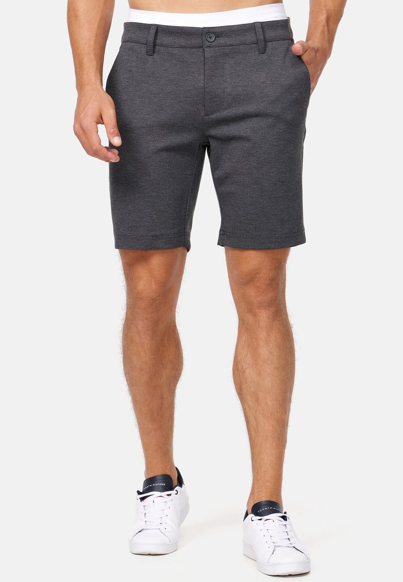 Homme portant un short gris foncé à boutons et des baskets blanches avec des touches noires, debout devant un fond uni.
