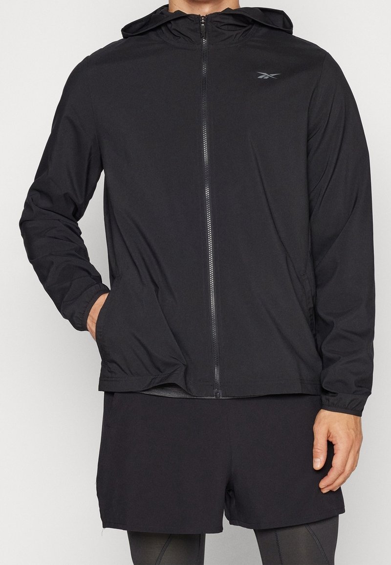 Homme portant une veste à capuche noire zippée avec le logo Reebok sur la poitrine et un short noir par-dessus un legging, main dans la poche de la veste.